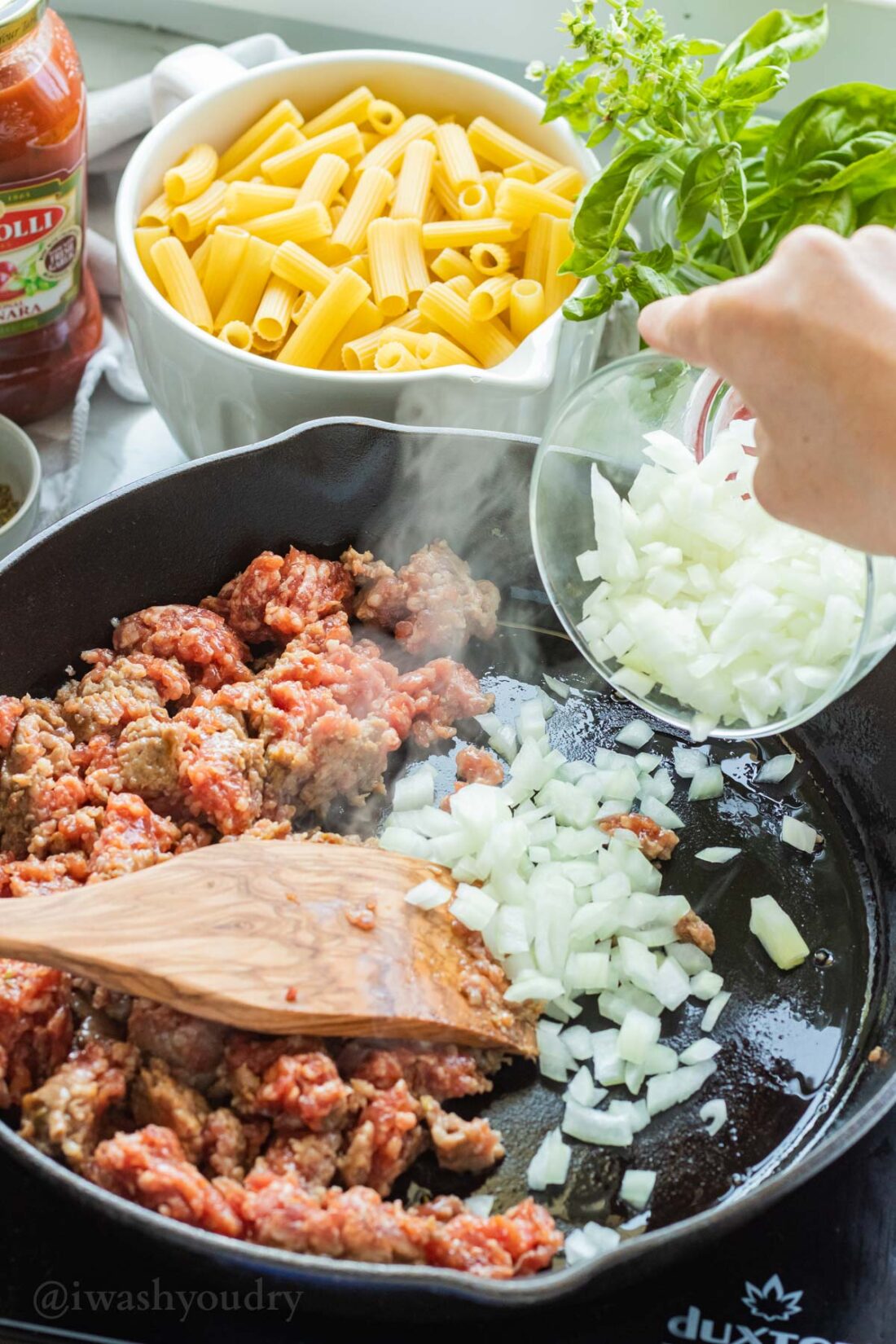 Raw italian sausage and onion in black frying pan with bowl of ziti noodles in background. 