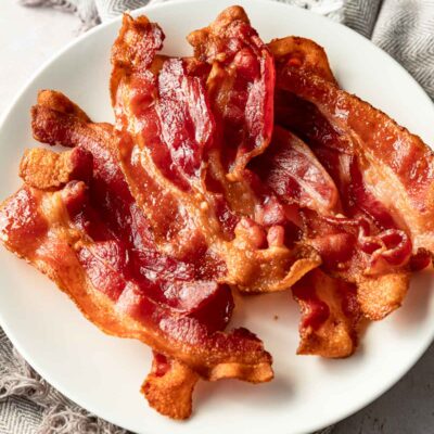 Cooked bacon slices on white plate with dish towel.