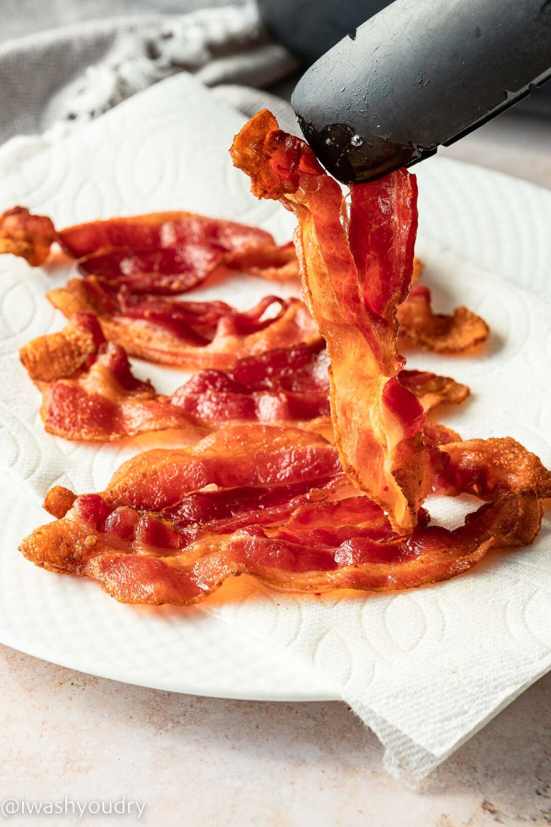 Tongs holding up cooked piece of air fryer bacon above paper towel. 