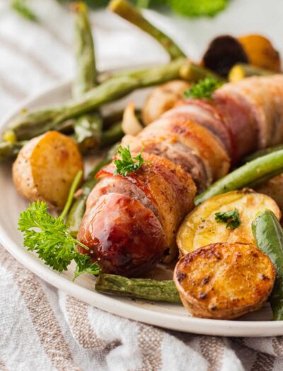 Cooked and sliced bacon wrapped bratwurst with potatoes and green beans on white plate.