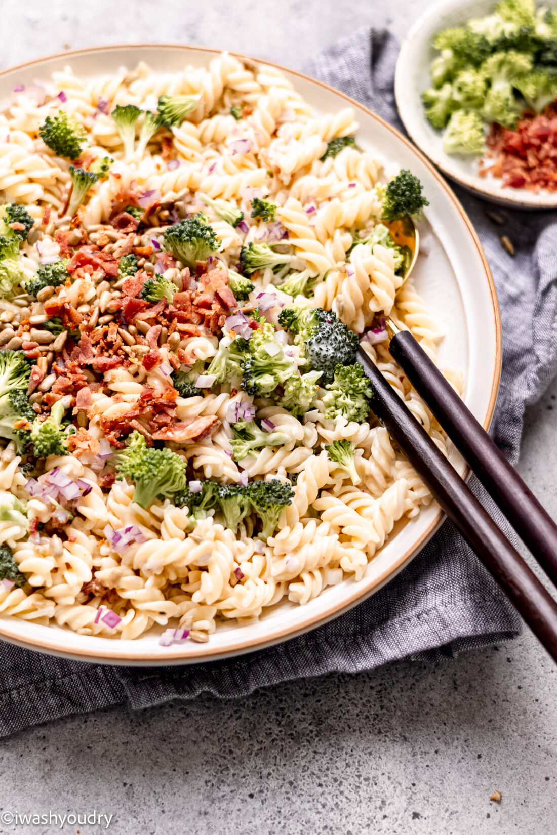 Bowl with creamy broccoli salad and bacon with serving utensils.