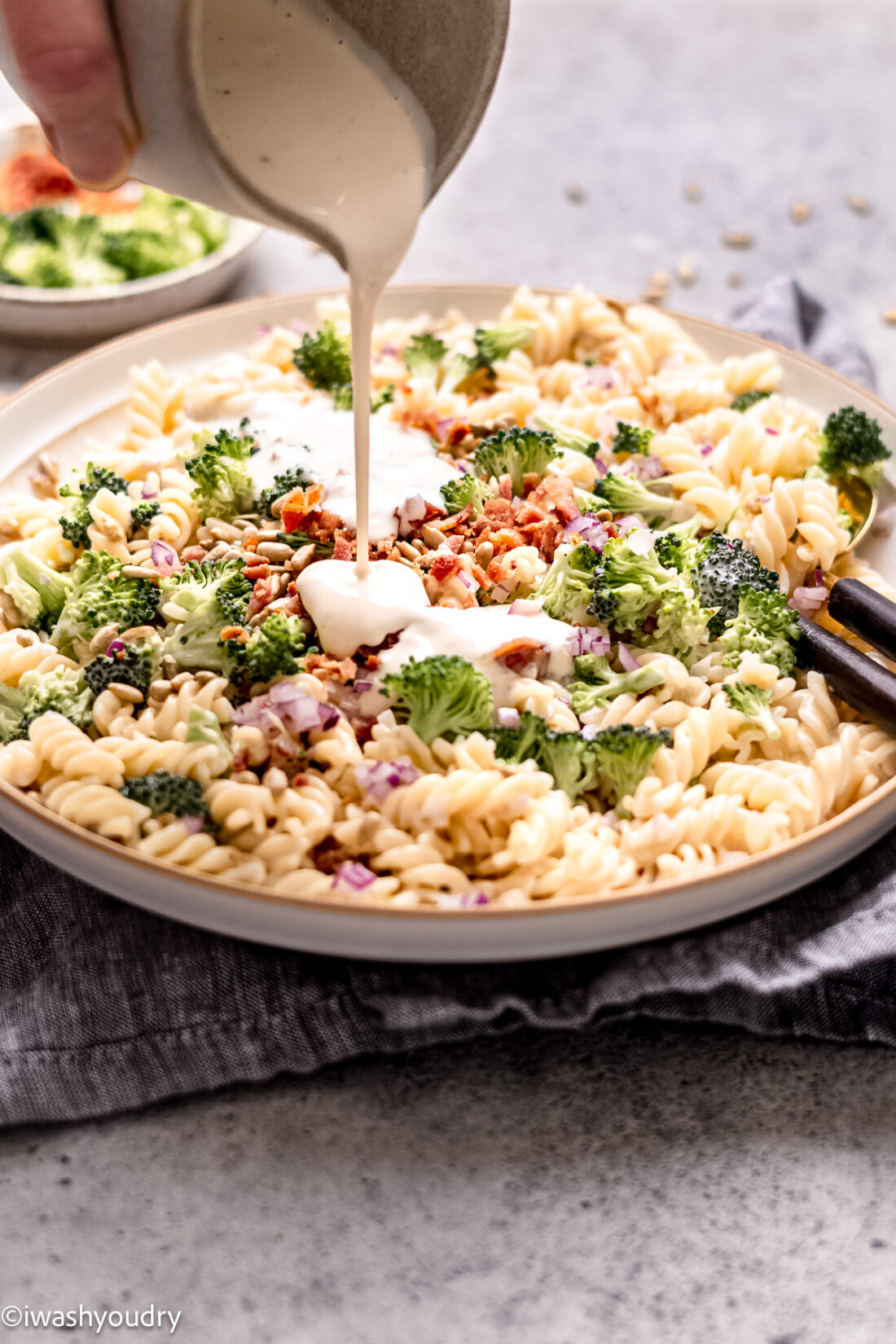 pouring white dressing over pasta salad with bacon.