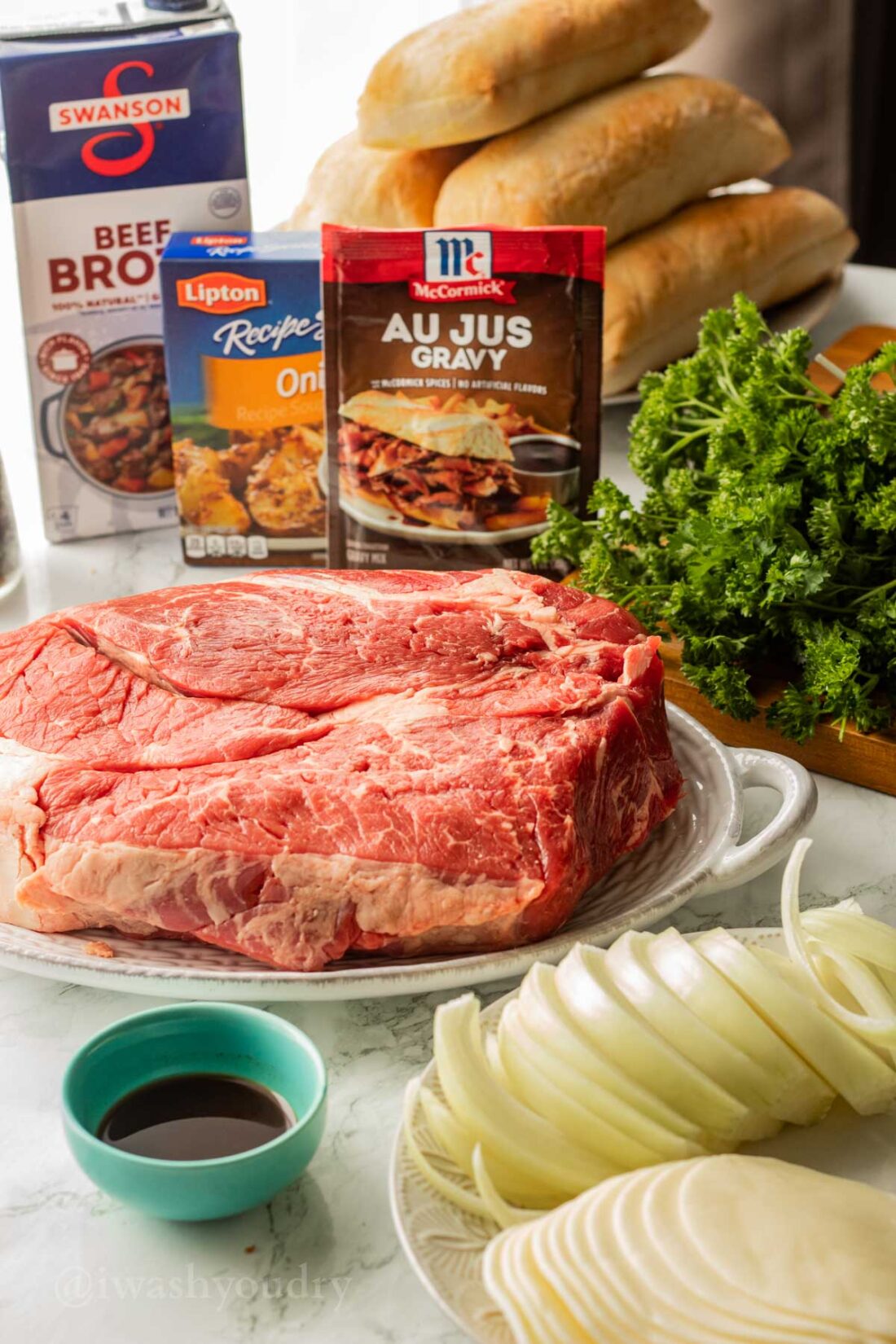 Ingredients for french dip sandwiches on white plates and blue dish on marble surface. 