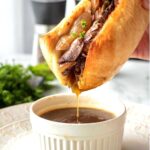 Cooked french dip sandwich dipping into white up of au jus.