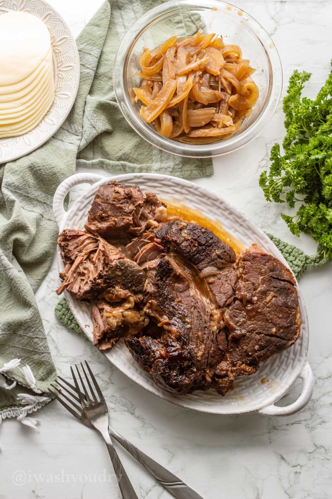 Cooked chuck roast on white platter with glass bowl of onions and forks on marble surface. 