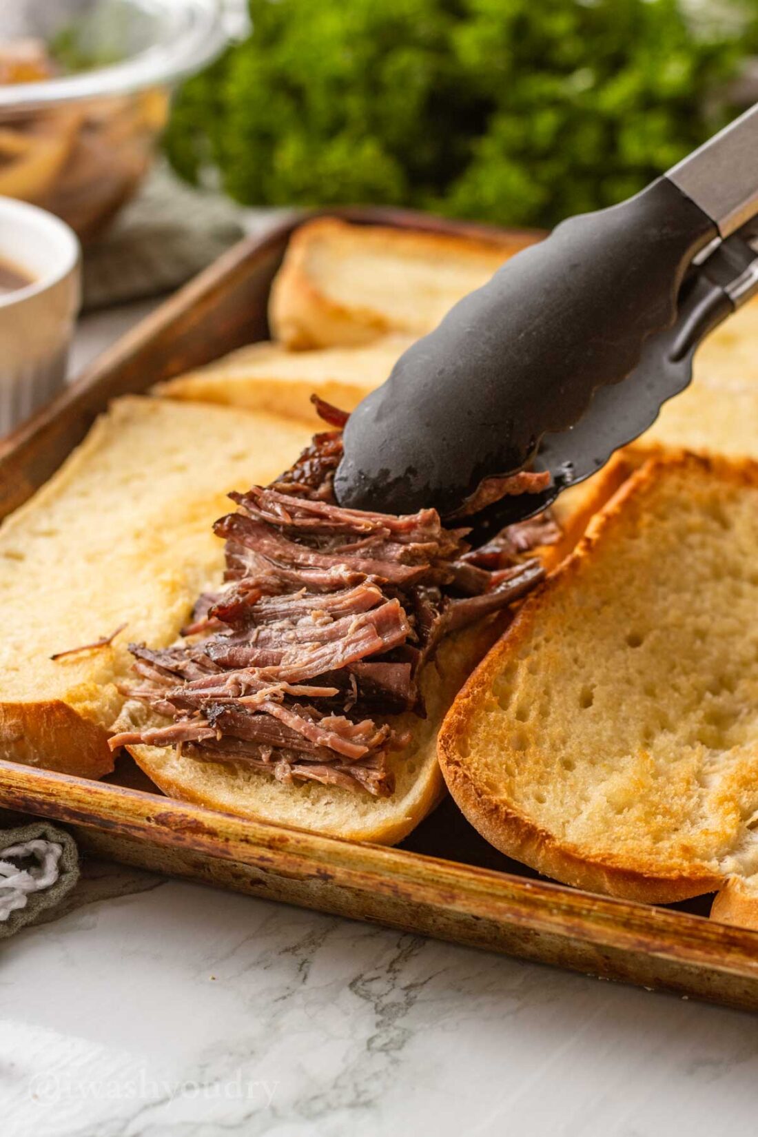 Black tongs placing shredded cooked beef on toasted buns. 