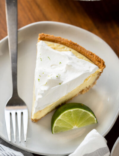 Slice of key lime pie on white plate with fork and lime slice.