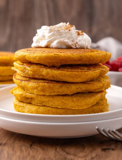 stack of pumpkin pancakes on white plate with whipped cream on top.