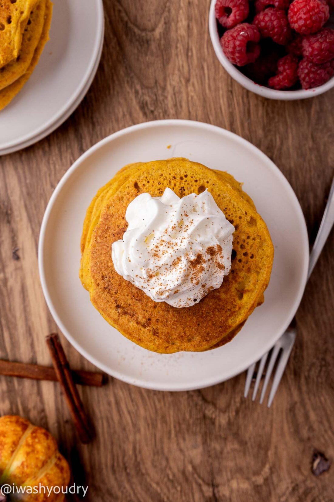 Stack of cooked pancakes on white plate near bowl of raspberries and cinnamon sticks. 