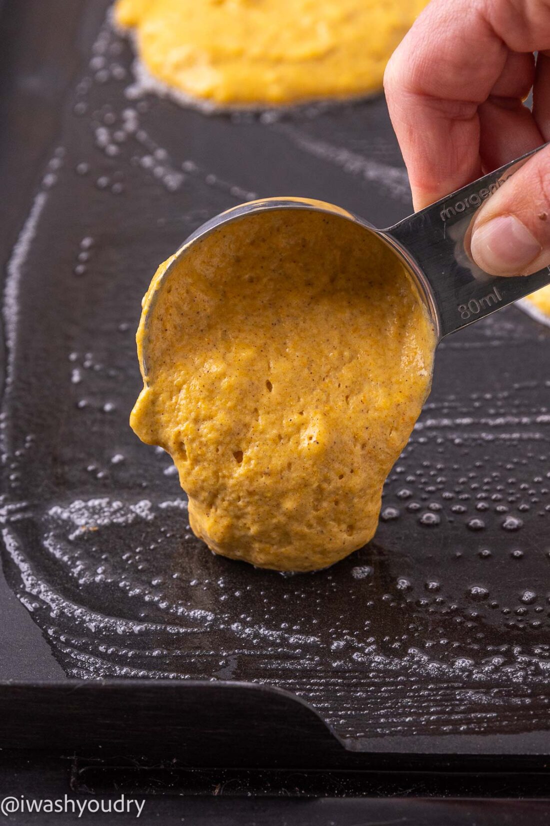 Cup pouring raw pumpkin pancake batter onto hot skillet. 