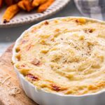 Baked roasted garlic dip in white dish over wood cutting board.