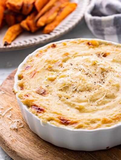 Baked roasted garlic dip in white dish over wood cutting board.