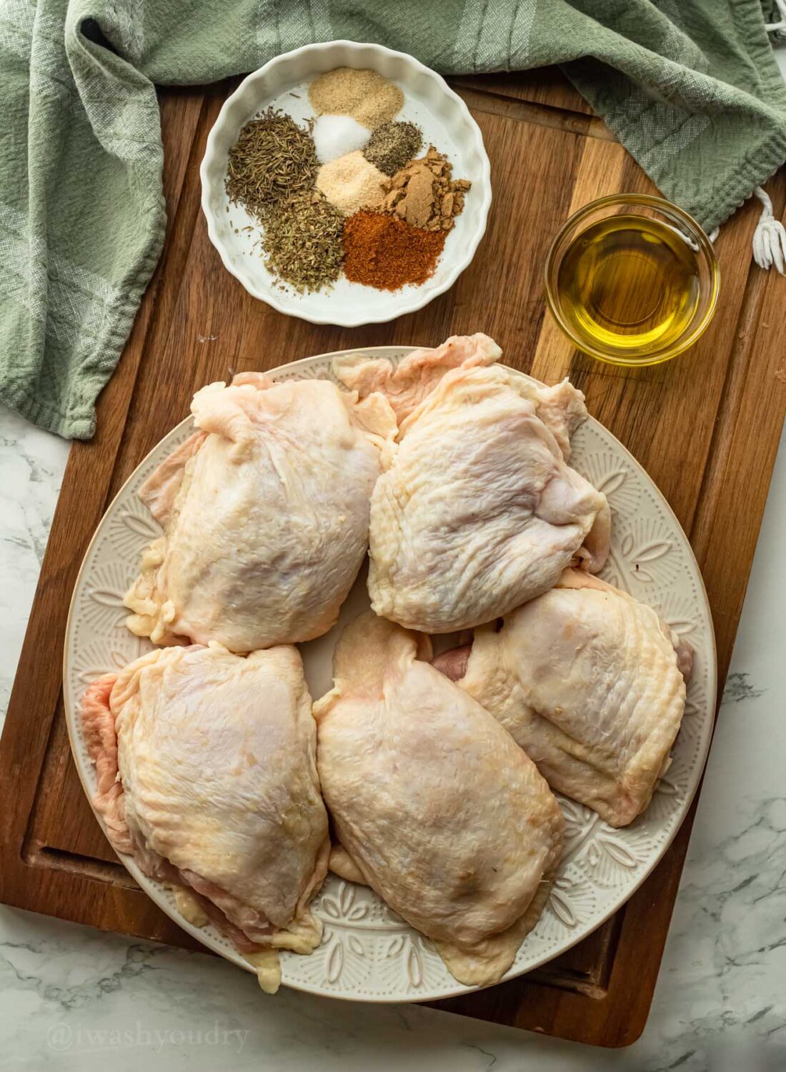 Raw chicken thighs o white plate with glass dishes of olive oil and spices on wooden cutting board. 