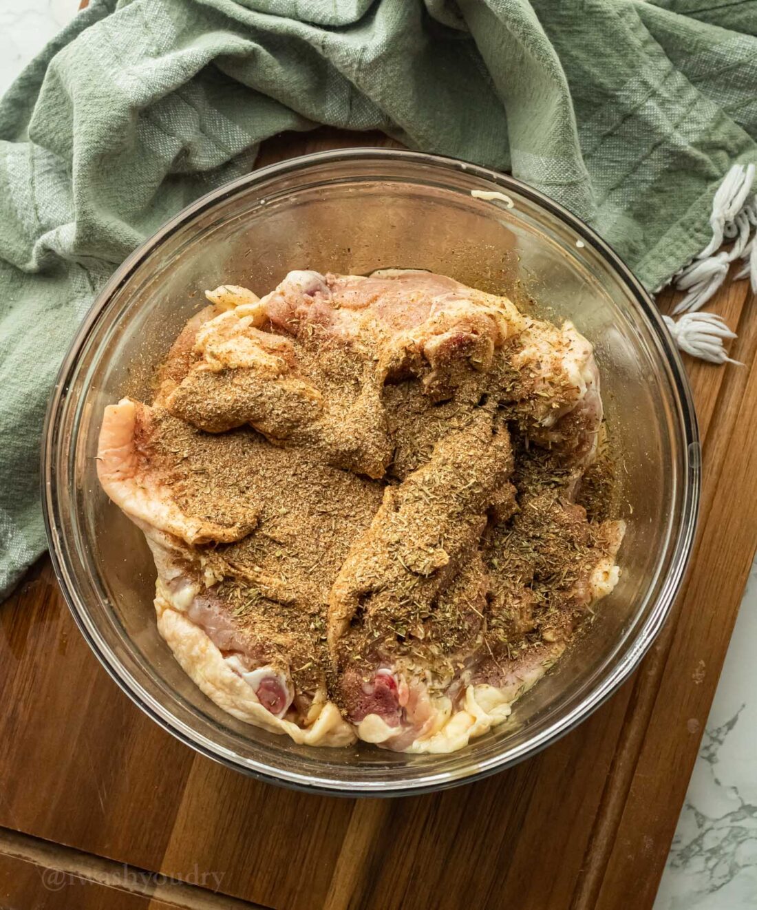 Raw chicken thighs in glass bowl with spices on top, resting on wood cutting board. 