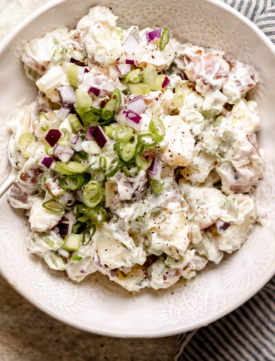 Creamy red potato salad in a white bowl with green onions and cracked pepper on top.