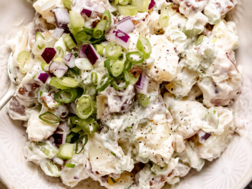 Creamy red potato salad in a white bowl with green onions and cracked pepper on top.