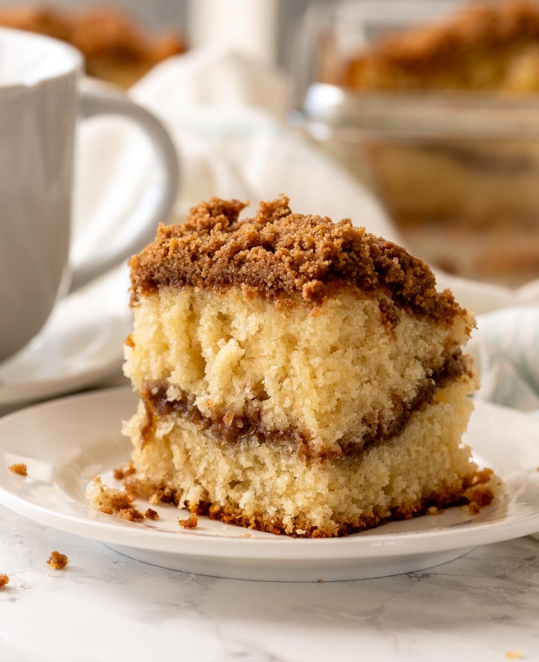 Slice of baked coffee cake on white plate near white mug. 