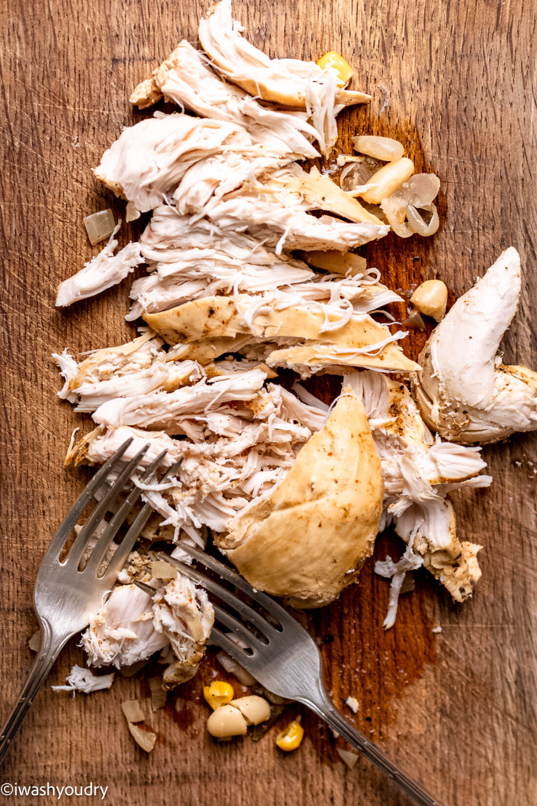 Forks shredding cooked chicken breasts on cutting board. 