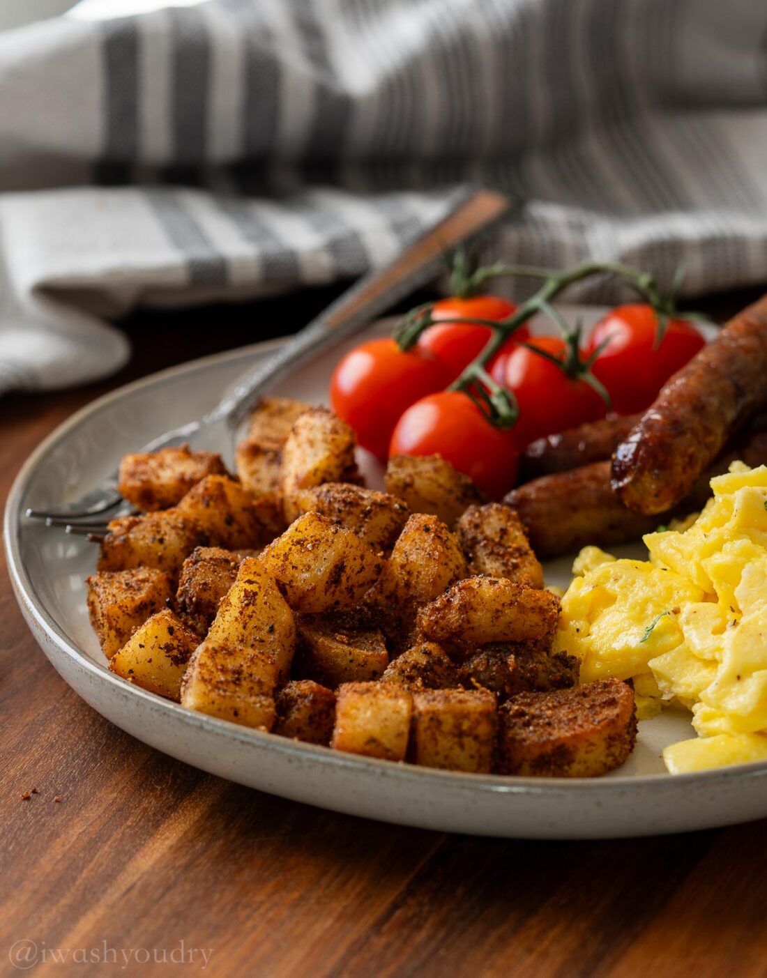 Cooked sausage, eggs, vine tomatoes, and breakfast potatoes on white plate. 