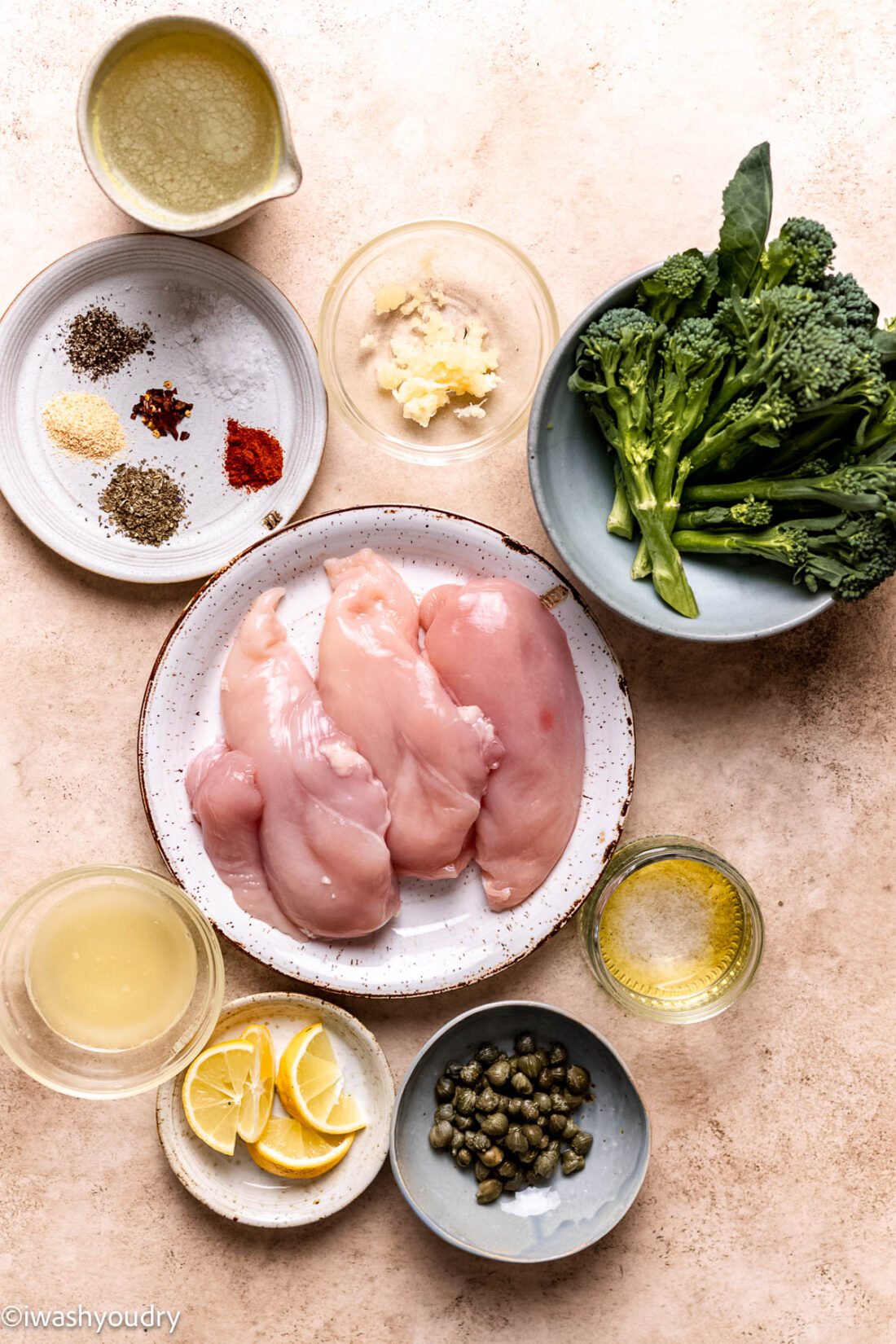 Ingredients for lemon chicken and broccolini in glass bowls and plates on marble countertop. 