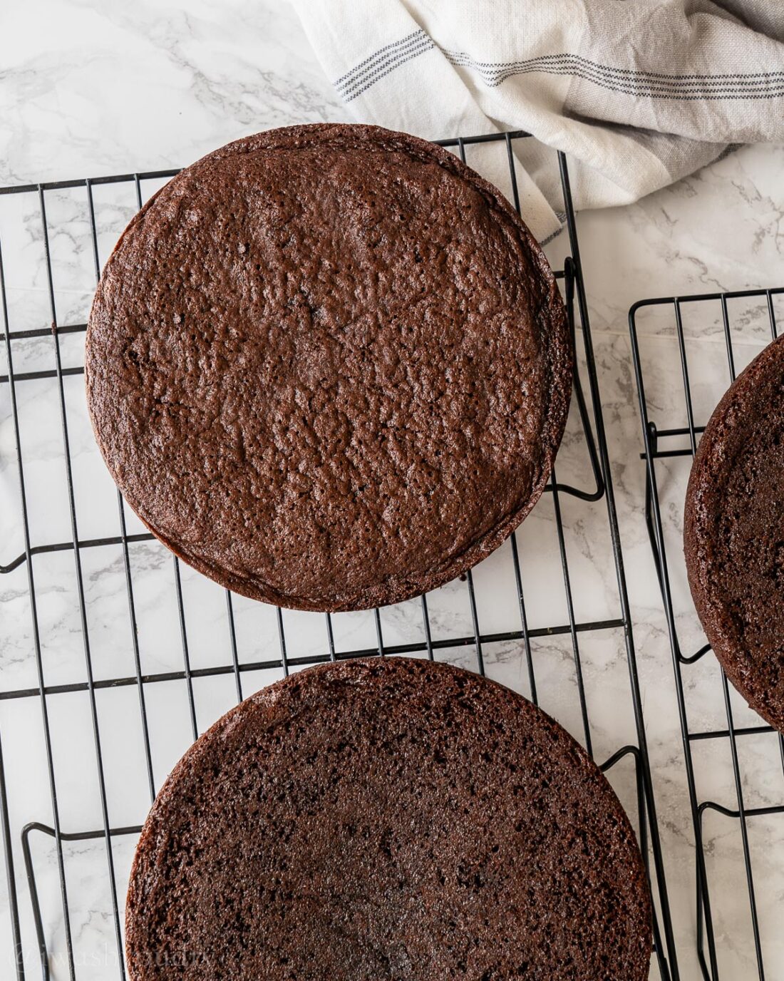 Chocolate cakes cooling on wire rack with white towel nearby.