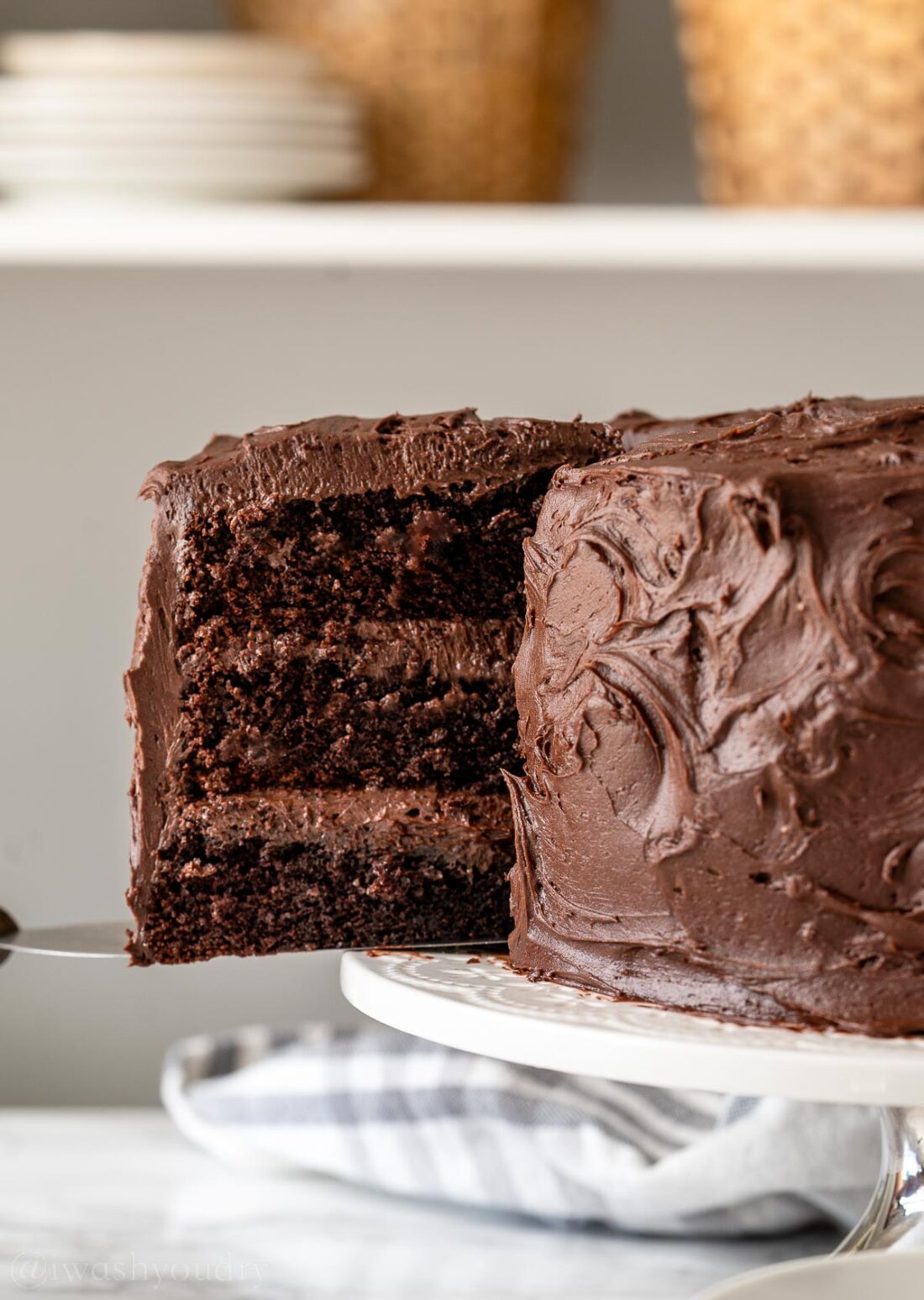 slice of triple layered chocolate cake with chocolate frosting being pulled out of a cake on a white stand.