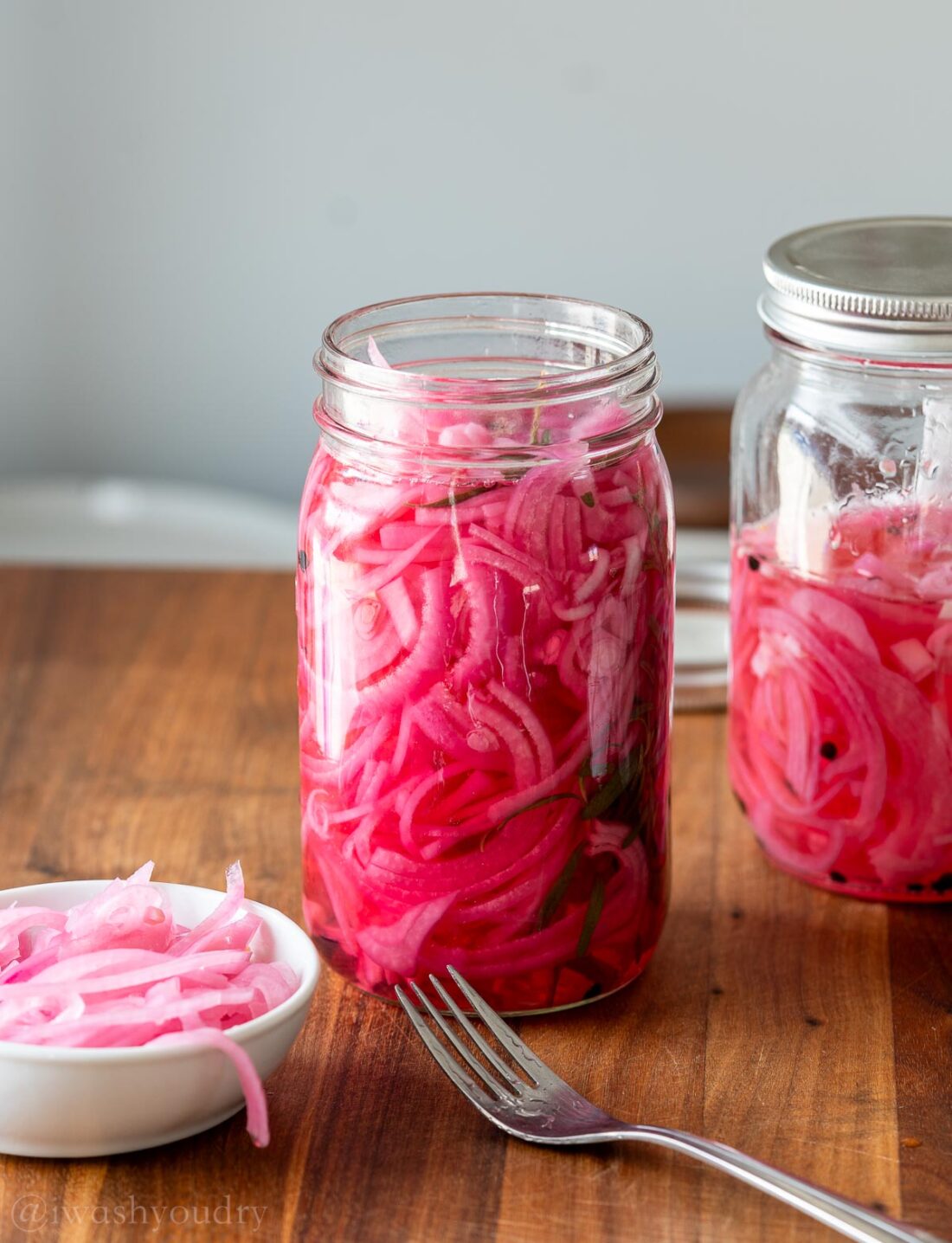 Jar of pickled onions on wood cuttin board near fork. 