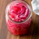 Pickled red onions in glass jar on wood cutting board.